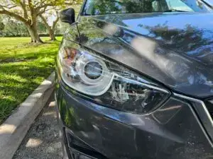 Close-up image of a like-new, shiny car headlight on a vehicle in Aventura, FL. The headlight lens is clear and polished, reflecting sunlight and the surrounding environment. The sleek, well-maintained appearance suggests recent cleaning or headlight restoration.