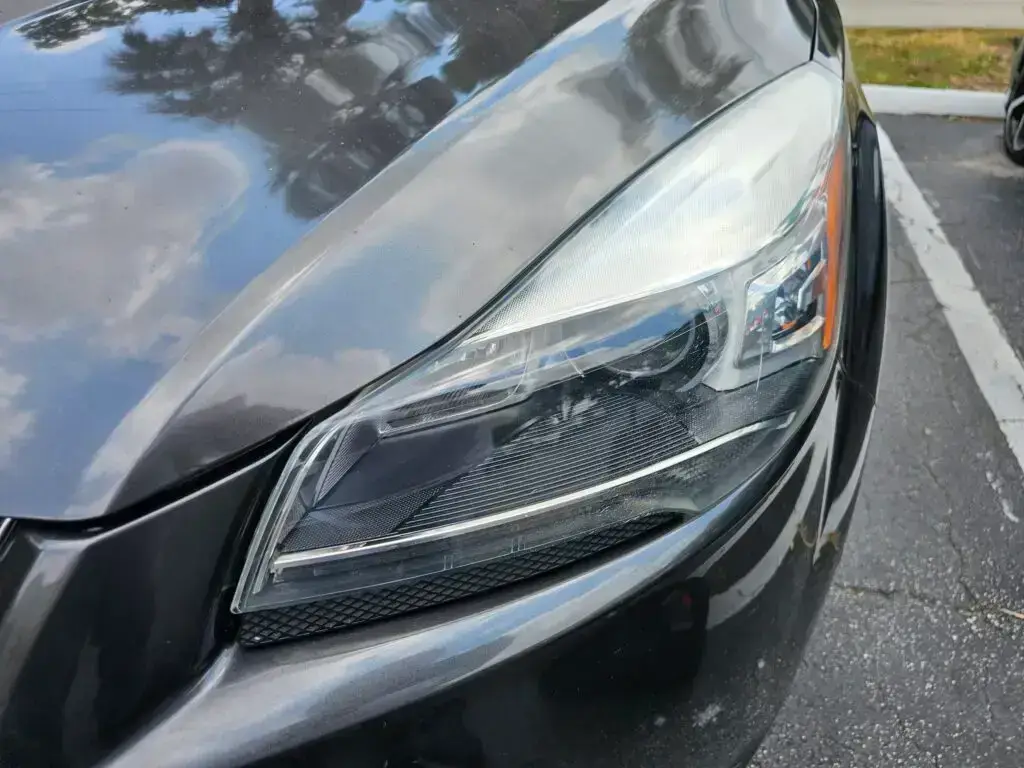 Close-up image of a like-new, shiny car headlight on a vehicle in Hallandale Beach, Florida. The headlight lens is clear and polished, reflecting sunlight and the surrounding environment. The sleek, well-maintained appearance suggests recent cleaning or headlight restoration.