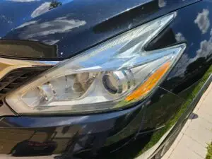 Close-up image of a like-new, shiny car headlight on a vehicle in Hialeah, Florida. The headlight lens is clear and polished, reflecting sunlight and the surrounding environment. The sleek, well-maintained appearance suggests recent cleaning or headlight restoration.