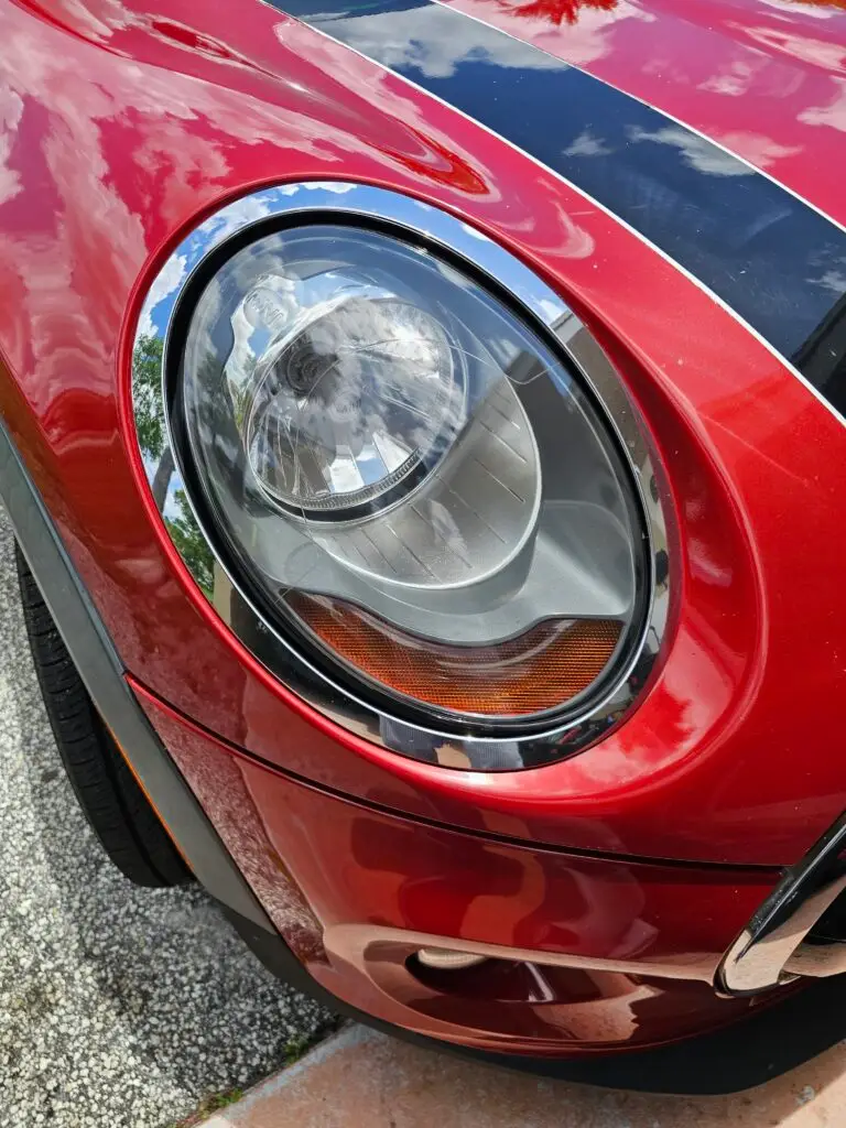 Close-up image of a like-new, shiny car headlight on a vehicle in Hollywood, Florida. The headlight lens is clear and polished, reflecting sunlight and the surrounding environment. The sleek, well-maintained appearance suggests recent cleaning or headlight restoration.