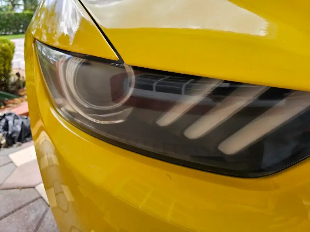 Close-up image of a like-new, shiny car headlight on a vehicle in Doral, Florida. The headlight lens is clear and polished, reflecting sunlight and the surrounding environment. The sleek, well-maintained appearance suggests recent cleaning or headlight restoration.