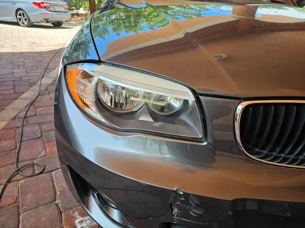 Close-up image of a like-new, shiny car headlight on a vehicle in Lauderdale by the sea, Florida. The headlight lens is clear and polished, reflecting sunlight and the surrounding environment. The sleek, well-maintained appearance suggests recent cleaning or headlight restoration.