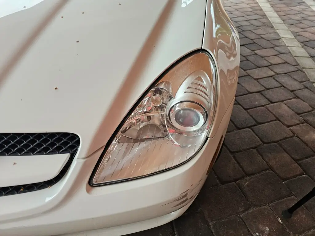 Close-up image of a like-new, shiny car headlight on a vehicle in Margate, Florida. The headlight lens is clear and polished, reflecting sunlight and the surrounding environment. The sleek, well-maintained appearance suggests recent cleaning or headlight restoration.