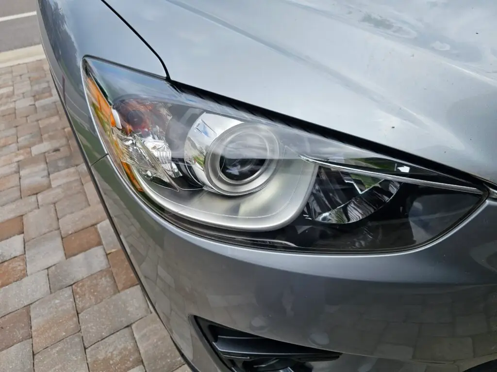 Close-up image of a like-new, shiny car headlight on a vehicle in Miami Gardens, Florida. The headlight lens is clear and polished, reflecting sunlight and the surrounding environment. The sleek, well-maintained appearance suggests recent cleaning or headlight restoration.