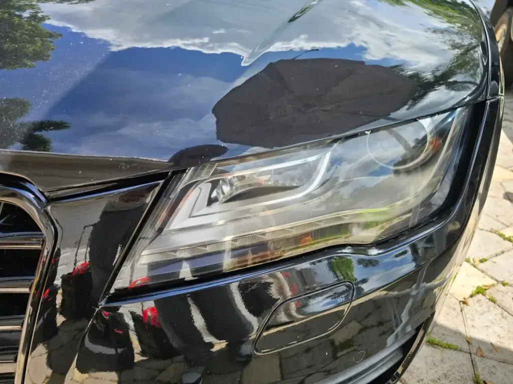 Close-up image of a like-new, shiny car headlight on a vehicle in Miami Lakes, Florida. The headlight lens is clear and polished, reflecting sunlight and the surrounding environment. The sleek, well-maintained appearance suggests recent cleaning or headlight restoration.