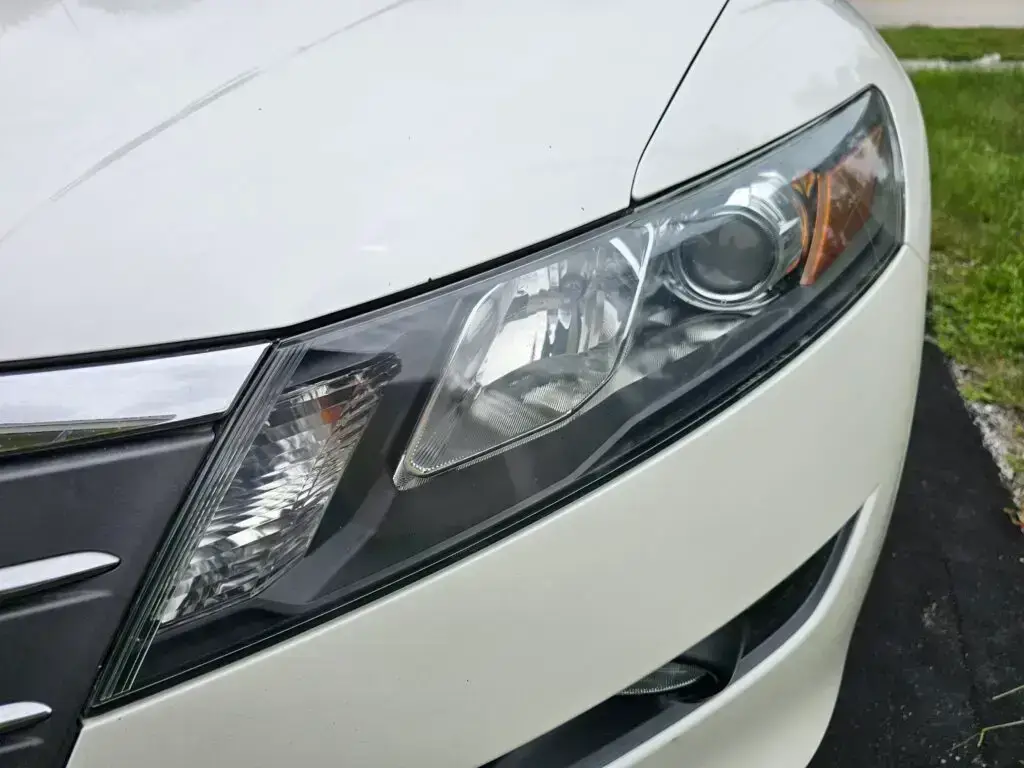 Close-up image of a like-new, shiny car headlight on a vehicle in Oakland Park, Florida. The headlight lens is clear and polished, reflecting sunlight and the surrounding environment. The sleek, well-maintained appearance suggests recent cleaning or headlight restoration.