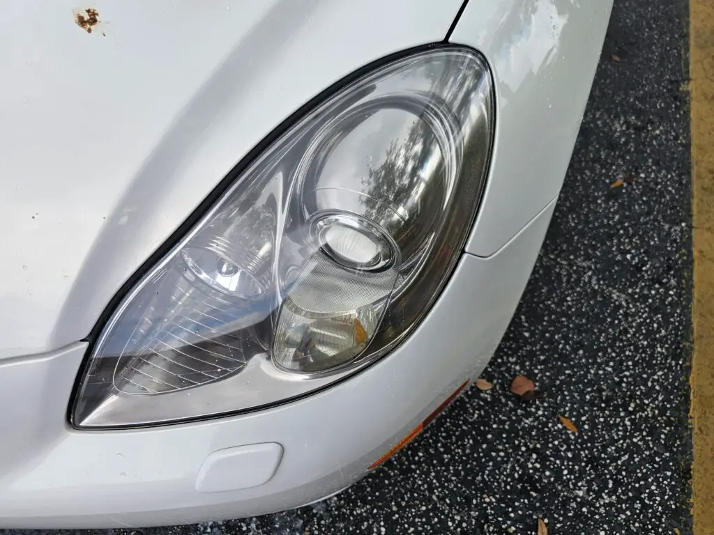Close-up image of a like-new, shiny car headlight on a vehicle in Plantation, Florida. The headlight lens is clear and polished, reflecting sunlight and the surrounding environment. The sleek, well-maintained appearance suggests recent cleaning or headlight restoration.