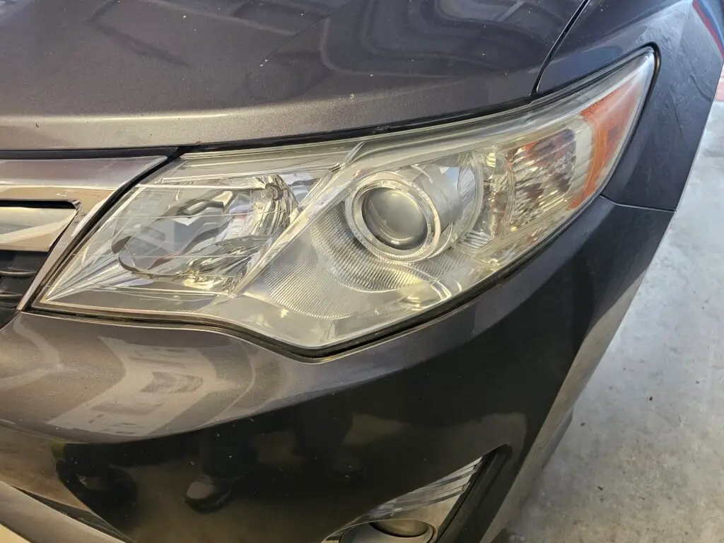Close-up image of a like-new, shiny car headlight on a vehicle in Pompano, Florida. The headlight lens is clear and polished, reflecting sunlight and the surrounding environment. The sleek, well-maintained appearance suggests recent cleaning or headlight restoration.