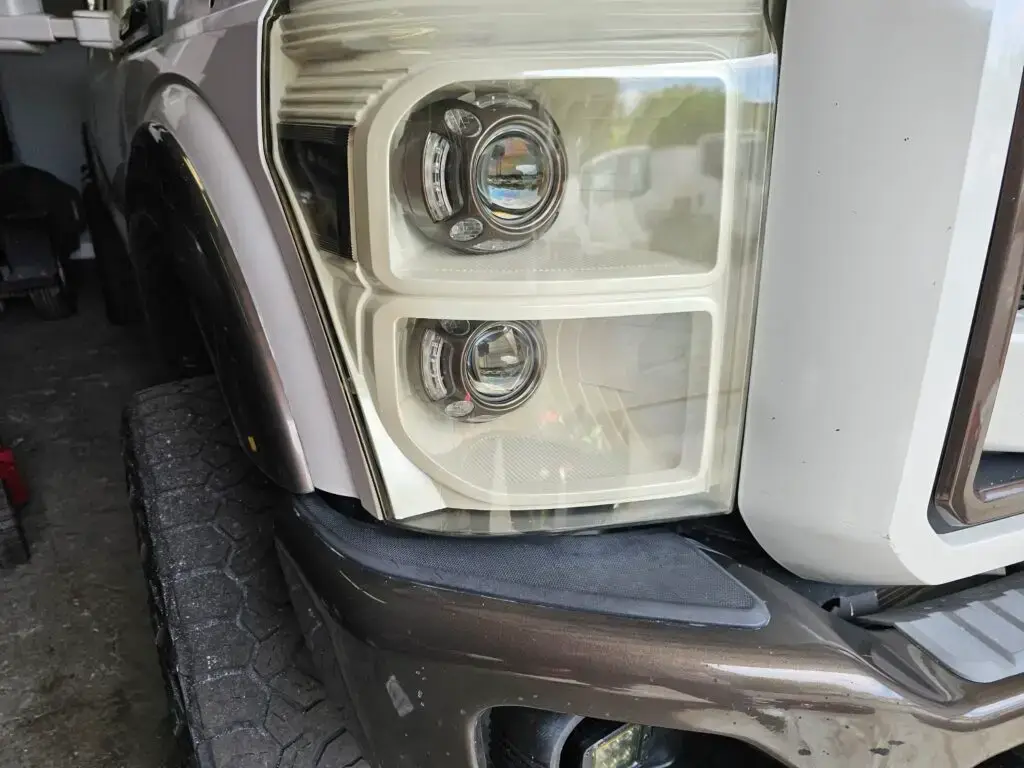 Close-up image of a like-new, shiny car headlight on a vehicle in Sunrise, Florida. The headlight lens is clear and polished, reflecting sunlight and the surrounding environment. The sleek, well-maintained appearance suggests recent cleaning or headlight restoration.