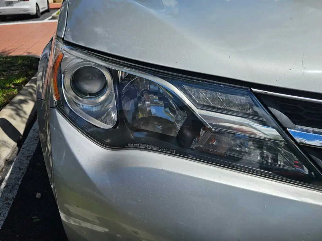 Close-up image of a like-new, shiny car headlight on a vehicle in West Park, Florida. The headlight lens is clear and polished, reflecting sunlight and the surrounding environment. The sleek, well-maintained appearance suggests recent cleaning or headlight restoration.