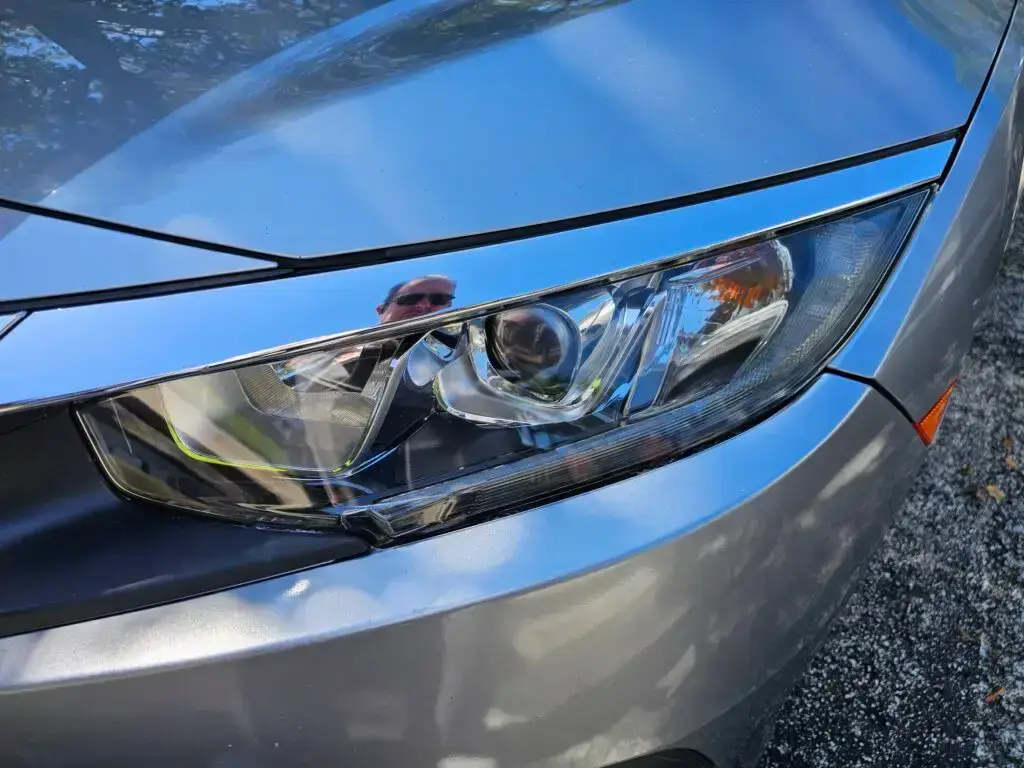 Close-up image of a like-new, shiny car headlight on a vehicle in Weston, Florida. The headlight lens is clear and polished, reflecting sunlight and the surrounding environment. The sleek, well-maintained appearance suggests recent cleaning or headlight restoration.