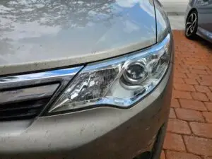 Close-up image of a like-new, shiny car headlight on a vehicle in coconut creek, Florida. The headlight lens is clear and polished, reflecting sunlight and the surrounding environment. The sleek, well-maintained appearance suggests recent cleaning or headlight restoration.