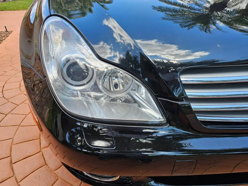 Close-up image of a like-new, shiny car headlight on a vehicle in Cooper City, Florida. The headlight lens is clear and polished, reflecting sunlight and the surrounding environment. The sleek, well-maintained appearance suggests recent cleaning or headlight restoration.