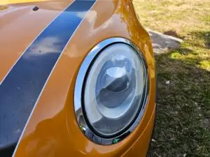 Close-up image of a like-new, shiny car headlight on a vehicle in coral springs, Florida. The headlight lens is clear and polished, reflecting sunlight and the surrounding environment. The sleek, well-maintained appearance suggests recent cleaning or headlight restoration.
