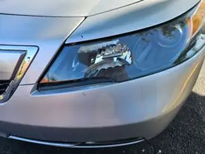 Close-up image of a like-new, shiny car headlight on a vehicle in Dania Beach, Florida. The headlight lens is clear and polished, reflecting sunlight and the surrounding environment. The sleek, well-maintained appearance suggests recent cleaning or headlight restoration.