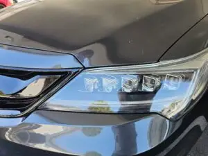 Close-up image of a like-new, shiny car headlight on a vehicle in Deerfield Beach, Florida. The headlight lens is clear and polished, reflecting sunlight and the surrounding environment. The sleek, well-maintained appearance suggests recent cleaning or headlight restoration.