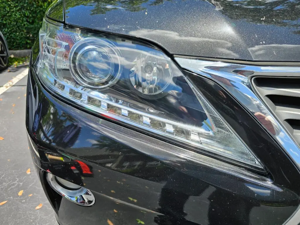 Close-up image of a like-new, shiny car headlight on a vehicle in Fort Lauderdale, Florida. The headlight lens is clear and polished, reflecting sunlight and the surrounding environment. The sleek, well-maintained appearance suggests recent cleaning or headlight restoration.