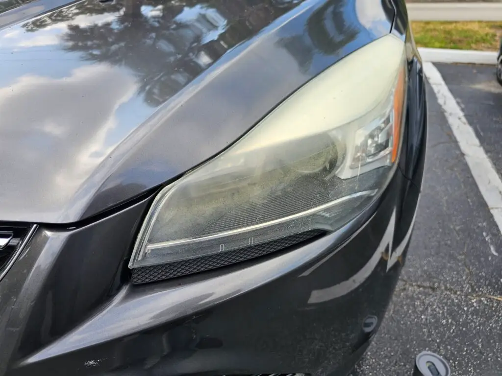 Close-up image of a faded and cloudy car headlight on a vehicle in Hallandale Beach, Florida. The headlight lens appears dull and foggy, likely due to oxidation or wear over time, and needs a headlight restoration.