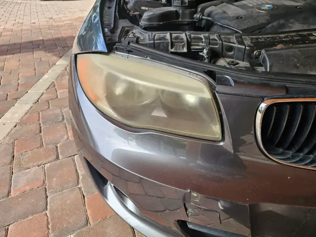 Close-up image of a faded and cloudy car headlight on a vehicle in Lauderdale by the sea, Florida. The headlight lens appears dull and foggy, likely due to oxidation or wear over time, and needs a headlight restoration.