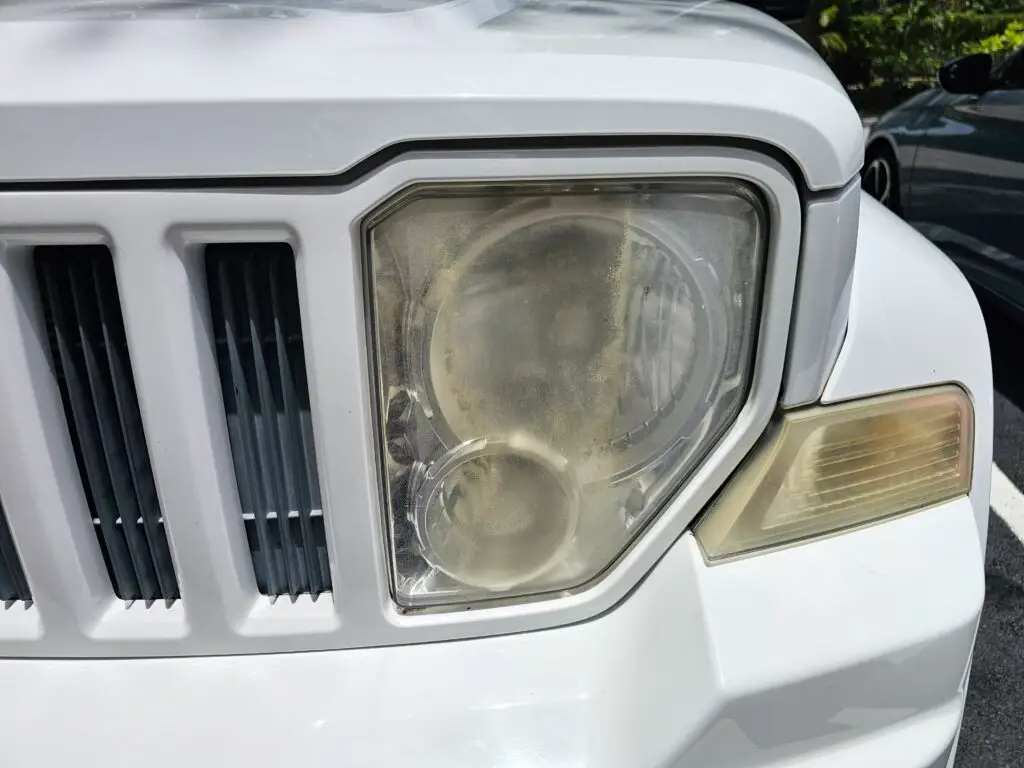 Close-up image of a faded and cloudy car headlight on a vehicle in Lauderdale Lakes, Florida. The headlight lens appears dull and foggy, likely due to oxidation or wear over time, and needs a headlight restoration.