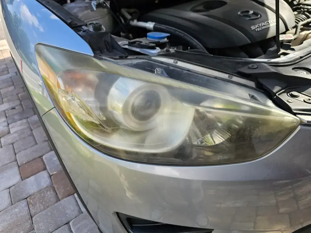 Close-up image of a faded and cloudy car headlight on a vehicle in Miami Gardens, Florida. The headlight lens appears dull and foggy, likely due to oxidation or wear over time, and needs a headlight restoration.