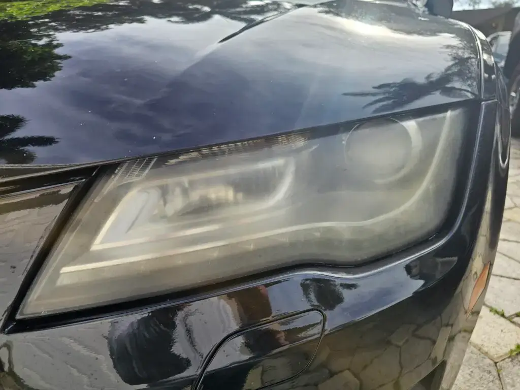 Close-up image of a faded and cloudy car headlight on a vehicle in Miami Lakes, Florida. The headlight lens appears dull and foggy, likely due to oxidation or wear over time, and needs a headlight restoration.