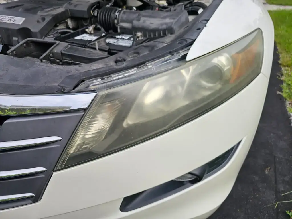 Close-up image of a faded and cloudy car headlight on a vehicle in Oakland Park, Florida. The headlight lens appears dull and foggy, likely due to oxidation or wear over time, and needs a headlight restoration.