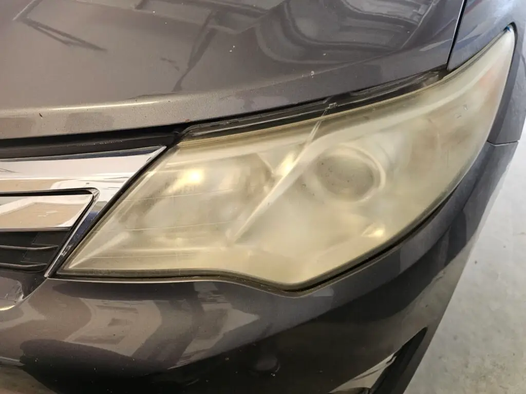 Close-up image of a faded and cloudy car headlight on a vehicle in Pompano, Florida. The headlight lens appears dull and foggy, likely due to oxidation or wear over time, and needs a headlight restoration.