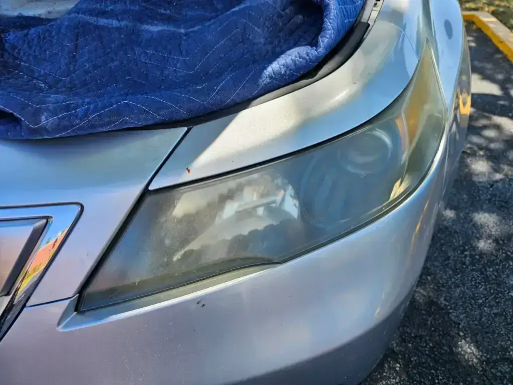 Close-up image of a faded and cloudy car headlight on a vehicle in Dania Beach, Florida. The headlight lens appears dull and foggy, likely due to oxidation or wear over time, and needs a headlight restoration