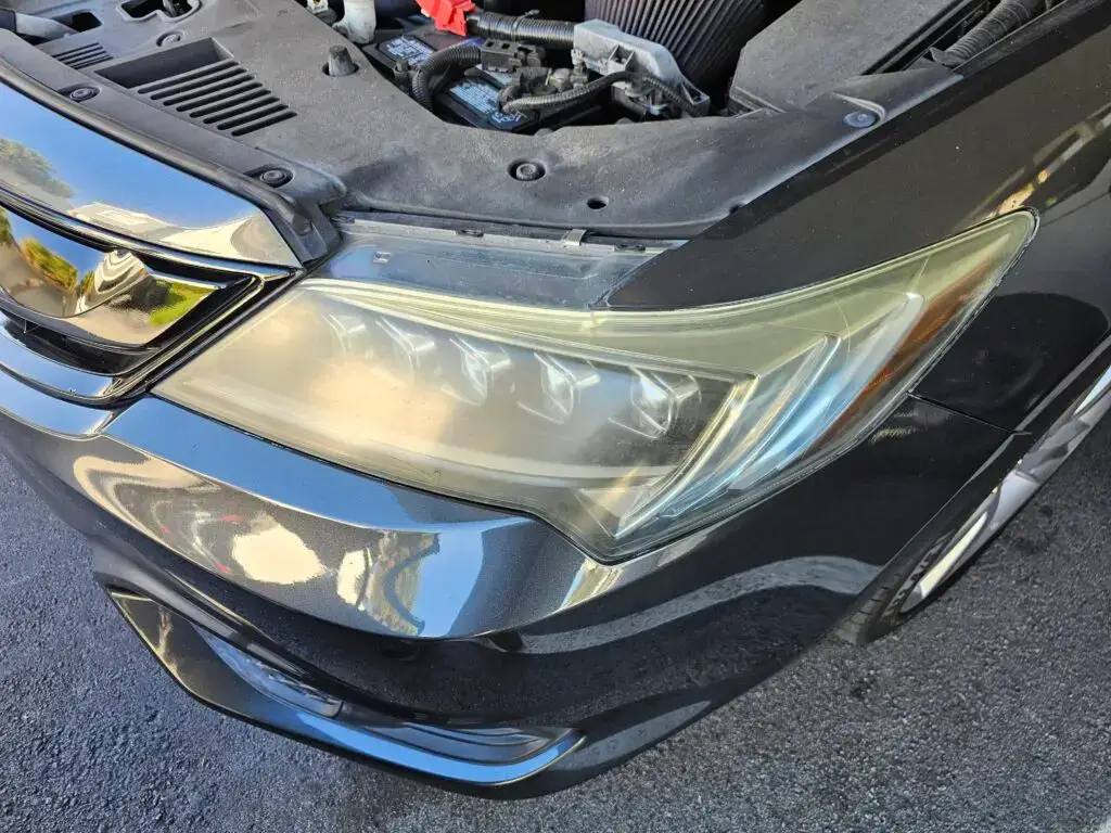 Close-up image of a faded and cloudy car headlight on a vehicle in Deerfield Beach, Florida. The headlight lens appears dull and foggy, likely due to oxidation or wear over time, and needs a headlight restoration.
